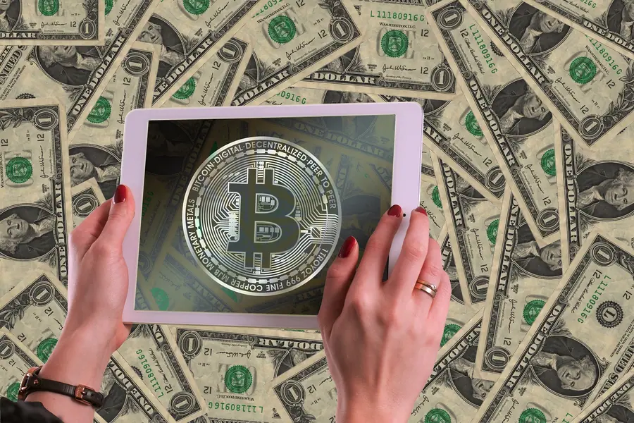 Hands holding a tablet showing a Bitcoin symbol over a background of banknotes
